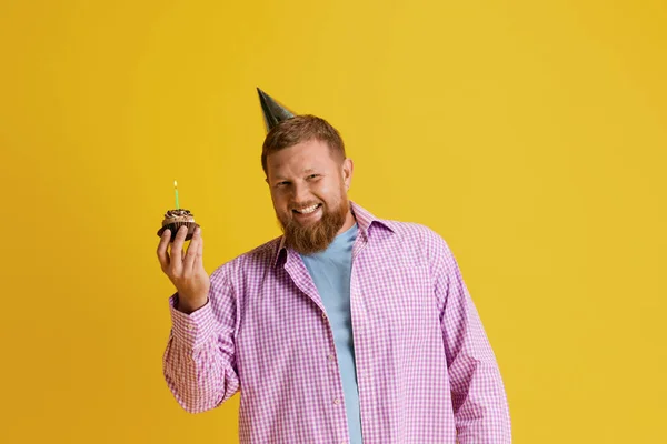 Happy, smiling, bearded man standing with cake, celebrating birthday against yellow studio background. Concept of human emotions, lifestyle, party, celebration, sales, fun, ad. Copy space for ad