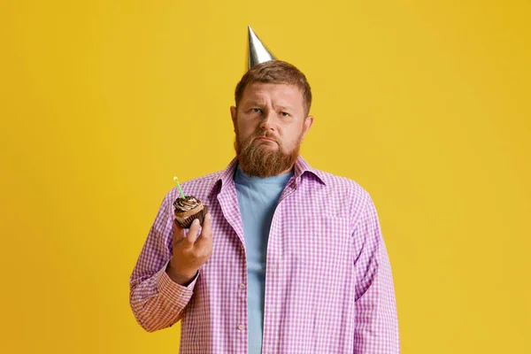Sad bearded man standing with cake, celebrating birthday against yellow studio background. Aging. Concept of human emotions, lifestyle, party, celebration, sales, fun, ad. Copy space for ad