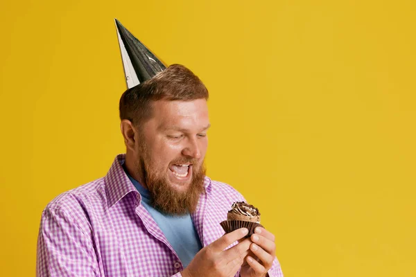 Excited bearded man feeling happy and positive, eating his birthday cupcake against over studio background. Concept of human emotions, lifestyle, party, celebration, sales, fun, ad. Copy space for ad