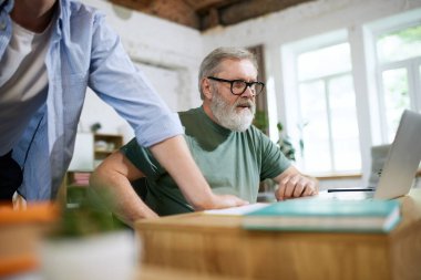 Yoğunlaşmış, üst düzey bir iş adamı ofiste iş arkadaşlarıyla birlikte çalışıyor, dizüstü bilgisayara bakıyor, sunum üzerinde çalışıyor. İş, eğitim, modernlik, yenilikler, yardım kavramı