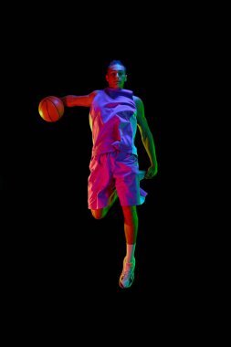 Galibiyet golü. Üniformalı genç adam basketbol oynuyor, neon ışıkta siyah stüdyonun arka planına top ile zıplıyor. Profesyonel spor, rekabet, hobi, oyun, rekabet kavramı