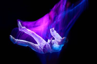 Gündelik giysiler içindeki genç adamın neon ışıklı siyah arka planda düşüşünün dinamik görüntüsü. Hareket kavramı, sanat, dans ve spor, moda, gençlik, reklam