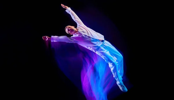 Young guy in sport style clothes in motion, doing somersaults against black studio background in neon with mixed lights effect. Concept of movements, art, dance and sport, fashion, youth, ad