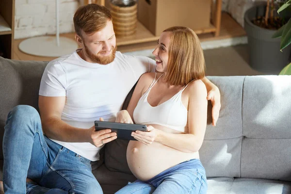 Young couple, future parents sitting at home and looking on tablet. Expecting baby, learning information. Concept of pregnancy, family, love, relationship, parenthood and childhood