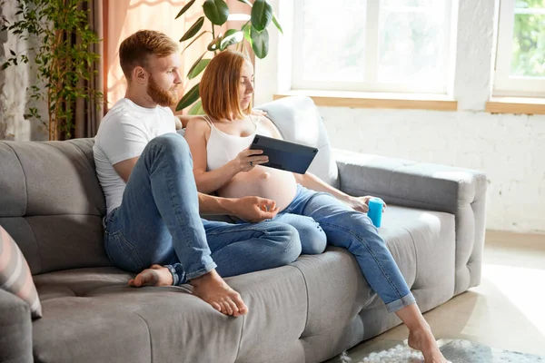 Young couple, future parents sitting at home and looking on tablet. Expecting baby, learning information. Concept of pregnancy, family, love, relationship, parenthood and childhood
