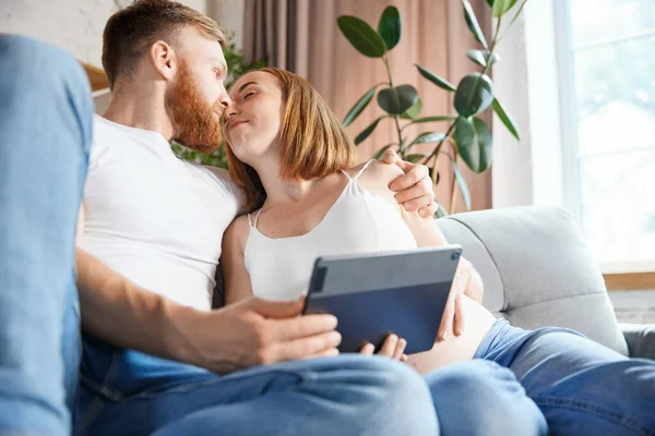 Kisses and love. Couple, parents sitting at home and looking on tablet. Expecting baby, learning information. Concept of pregnancy, family, love, relationship, parenthood and childhood
