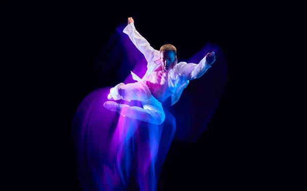 Dynamic image of stylish young man in white clothes humping against black studio background in neon with mixed lights effect. Concept of movements, art, dance and sport, fashion, youth, ad