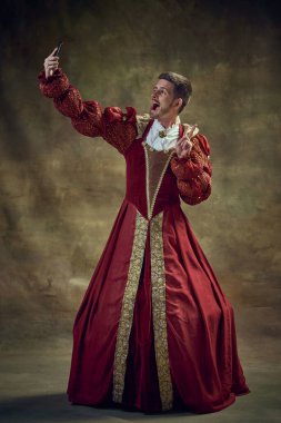 Genç bir adam, orta çağ kraliyet mensubu kadın kıyafeti giymiş koyu yeşil arka planda cep telefonuyla selfie çekiyor. Tarihi retrospektifler, moda, kışkırtıcı projeler, sosyal medya kavramı