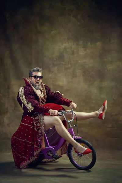 Man, medieval royal person in female dress and shoes riding little bike over vintage background. Humorous emotions. Concept of historical retrospectives, fashion, provoking projects, gender fluidity