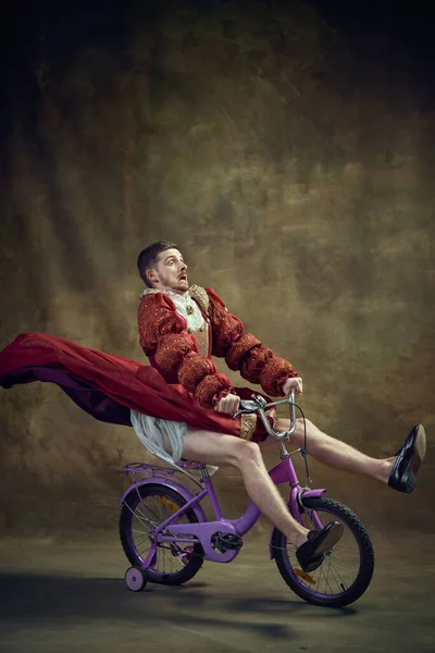 Young man, medieval royal person in female dress riding little bike on vintage background. Humorous emotions. Speed. Concept of historical retrospectives, fashion, provoking projects, gender fluidity