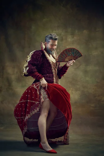 Drama. Humorous representation of medieval royal person, man in female dress with fan over vintage green background. Concept of historical retrospectives, fashion, provoking projects, gender fluidity