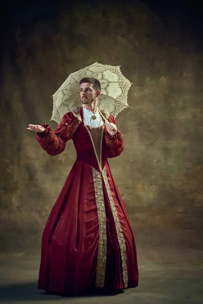 Historical royalplay. Medieval prince, nobleman in female dress, holding umbrella against vintage green background. Concept of historical retrospectives, fashion, provoking projects, gender fluidity