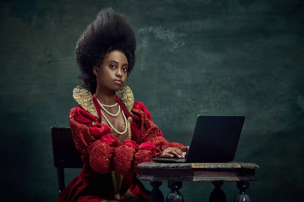 中世纪的公主 年轻的非洲女子穿着优雅的红色服装在笔记本电脑上工作与老式绿色背景 美与时尚的概念 广告的比较 — 图库照片