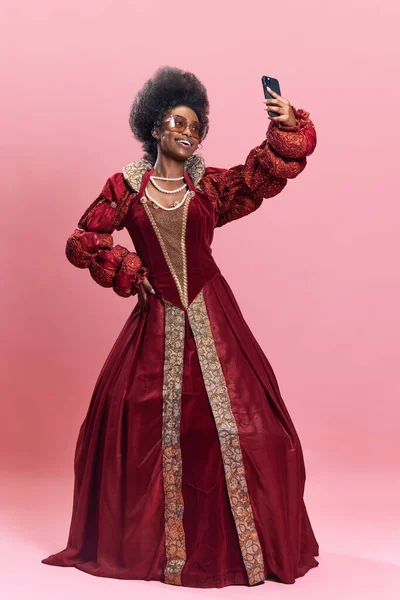非洲的年轻女子 身穿红衣服的公主 戴着太阳镜 手持手机 背景是粉色的工作室 有影响力的人历史 美与时尚的概念 时代的比较 — 图库照片