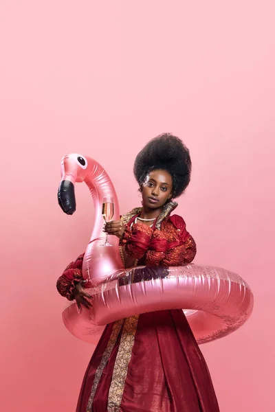 美丽的非洲女人 以公主的形象出现在游泳圈中 在粉色工作室的背景下举起香槟 美与时尚的概念 时代的比较 — 图库照片