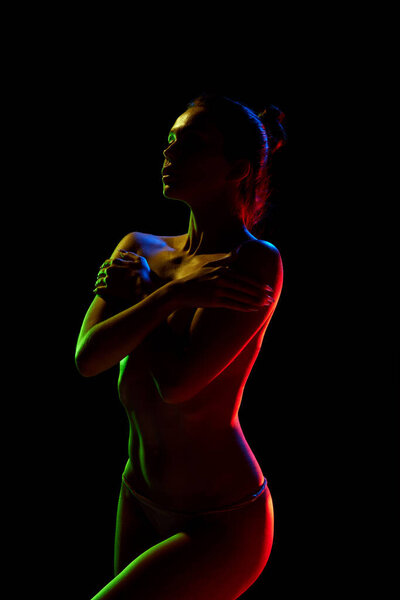 Tender woman with slim, naked body covering breast with hands against black studio background in neon lights. Concept of female beauty, body art, aesthetics, femininity and sensuality.