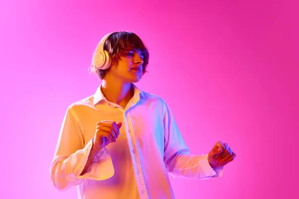 Young guy in white shirt listening to music in headphones and dancing against pink studio background in neon lights. Concept of human emotions, facial expression, youth, lifestyle. Ad