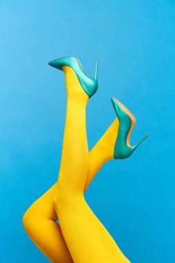 Şık, topuklu, yeşil ayakkabılı ve mavi arka planda sarı taytlı kadın bacakları. Renkli fotoğrafçılık. Moda anlayışı, yaratıcılık, hayal gücü. İlan için alanı kopyala