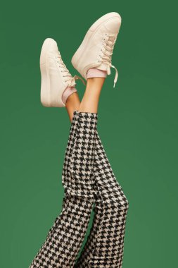 Beyaz spor ayakkabılı kadın bacakları ve yeşil arka planda kareli pantolonlar. Zeki, sıradan, iş tarzı. Renkli fotoğrafçılık. Moda anlayışı, yaratıcılık, hayal gücü. İlan için alanı kopyala