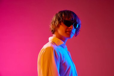 Beyaz tişörtlü ve güneş gözlüklü genç bir adamın portresi. Pembe stüdyonun arka planında neon ışıklarla poz veriyor. İnsan duyguları, yüz ifadesi, gençlik, yaşam tarzı kavramı. Ad