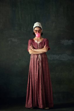 Ortaçağ hizmetçisinin portresi, tarihi kıyafetler giyiyor ve eski yeşil arka planda sakız yiyor. Tarih kavramı, çağların karşılaştırması, güzellik, sanat, yaratıcılık