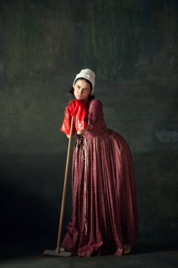 Sıkıldım ve yoruldum. Güzel kadın, ortaçağ elbiseli hizmetçi paspasa yaslanmış büyük kırmızı lastik eldivenlerle klasik yeşil arka planda. Tarih kavramı, çağların karşılaştırması, güzellik, sanat, yaratıcılık