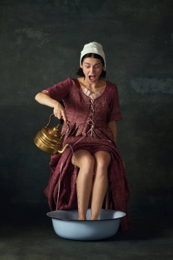 Ortaçağ kadınının görüntüsü. Sandalyeye oturmuş, bacakları kaseye girmiş ve eski yeşil arka plana sıcak su döküyor. Tarih kavramı, çağların karşılaştırması, güzellik, sanat, yaratıcılık