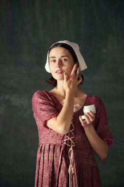 Ortaçağ hizmetçisi görünümündeki güzel genç bir kadın yüzüne krem sürüyor. Eski yeşil arka plana karşı. Cilt bakımı. Tarih kavramı, dönemlerin karşılaştırılması, güzellik, sanat, yaratıcılık, kozmetoloji