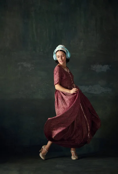 Medieval woman in elegant historical dress, maid listening to music in headphone and dancing against vintage green background. Concept of history, comparison of eras, beauty, art, creativity