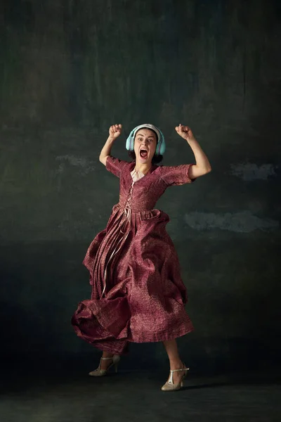 Emotional woman in elegant renaissance dress, medieval maid listening to music in headphone and dancing over vintage green background. Concept of history, comparison of eras, beauty, art, creativity
