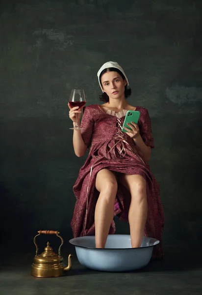 Relaxation. Woman in renaissance costume, maid sitting with legs into bowl, drinking wine and looking on phone against vintage green background. Concept of history, comparison of eras, art, creativity