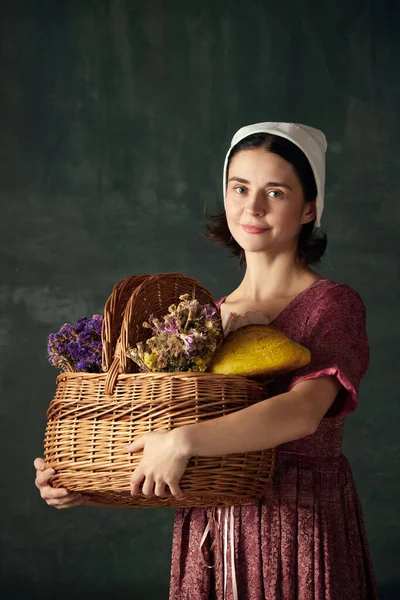 Countryside aesthetics. Elegant young woman, medieval maid standing with basket with flowers and bread against vintage green background. Concept of history, comparison of eras, beauty, art, creativity
