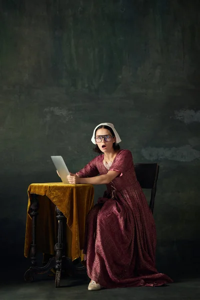 Emotional woman in renaissance dress, maid sitting at table with tablet and looking with shock against vintage green background. Concept of history, comparison of eras, beauty, art, creativity