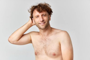 Gri stüdyo geçmişine bakarak gülümseyen kıvırcık saçlı, yakışıklı olgun bir adamın portresi. Hazırlanıyor. Erkeklerin güzellik anlayışı, vücut ve cilt bakımı, kozmetoloji, sağlık, reklam