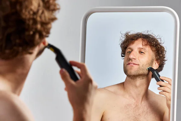 成熟したシャツレスの男性は 鏡を見て灰色のスタジオの背景に対してカミソリで剃ります 朝の準備をする メンズビューティー ボディ スキンケア 化粧品 ウェルネス 広告のコンセプト — ストック写真