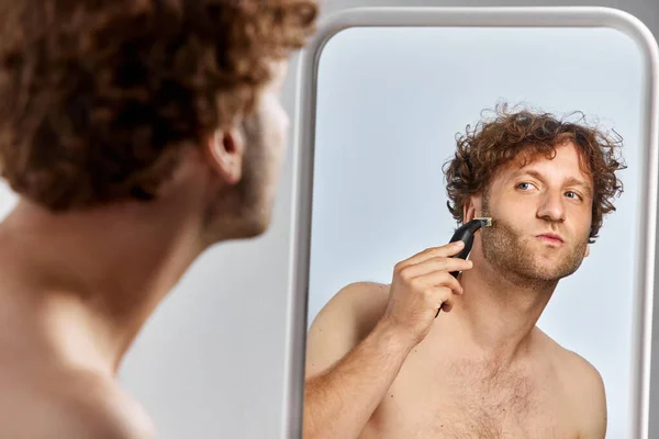 Mature shirtless man looking in mirror and shaving with razor against grey studio background. Getting ready in the morning. Concept of mens beauty, body and skin care, cosmetology, wellness, ad