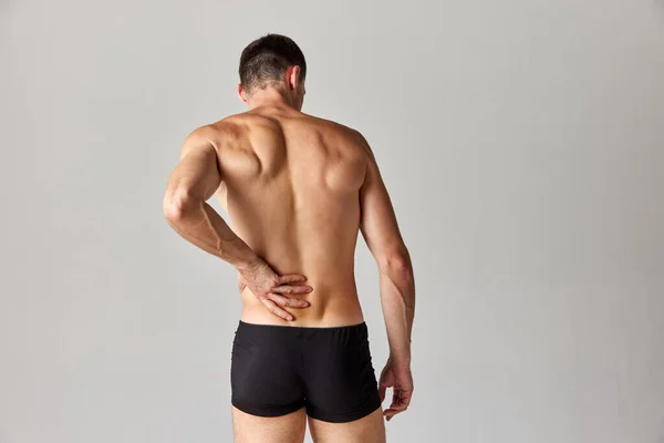 Back pains. Muscular man standing in underwear and holding his back against grey studio background. Medical treatment. Concept of mens health and beauty, body care, fitness, wellness, ad