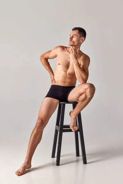 Full-length portrait of handsome, muscular young man sitting on chair in underwear and posing against grey studio background. Concept of mens health and beauty, body care, fitness, wellness, ad