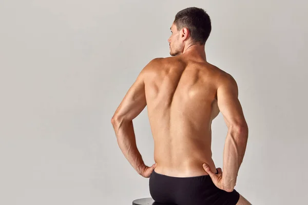 Back view image of muscular young shirtless man with relief, sign back sitting in underwear against grey studio background. Concept of mens health and beauty, body care, fitness, wellness, ad