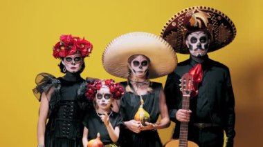 Şşş. Sessiz olun. Erkek, kadın ve kızlar, sarı arka planda şeker kafatası makyajı ve kostümler yapıyorlar. Cadılar Bayramı kavramı, El Dia de Muertos, bayramlar ve festivaller, Meksika gelenekleri, aile