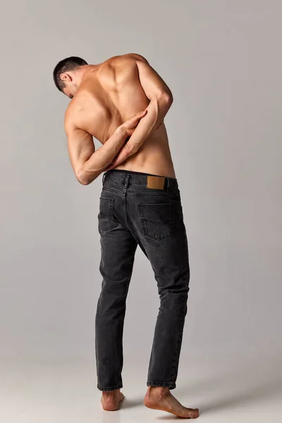 Young man with muscular, strong, relief body, back posing shirtless in jeans against grey studio background. Concept of mens health and beauty, body care, fitness, wellness, ad