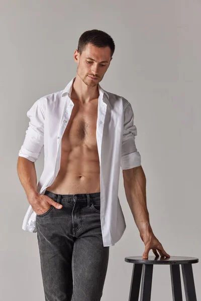 Handsome young man with muscular, relief body posing in white shirt and jeans against grey studio background. Concept of mens health and beauty, fashion, body care, fitness, wellness, ad