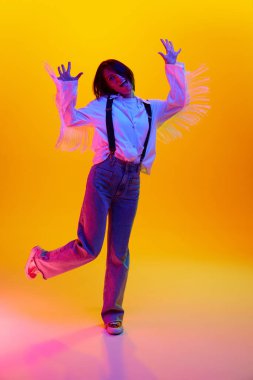 30 'lu yaşlarda, turuncu stüdyonun arka planında neon ışıklarıyla neşeli bir şekilde zıplayan pozitif kadının tam boy görüntüsü. İnsan duyguları kavramı, moda, yaşam tarzı, yüz ifadesi, reklam