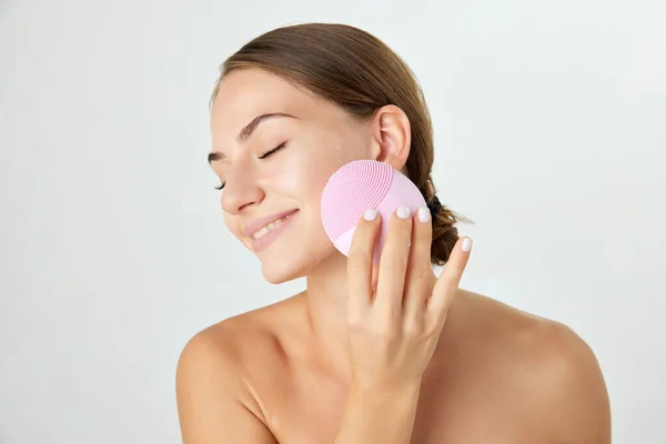 Portrait of beautiful young woman with well-kept using cleansing silicone brush isolated on white background. Concept of natural beauty, cosmetology and cosmetics, skin care, spa. Ad