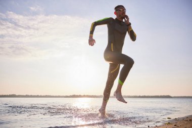 Profesyonel triatlet, dalgıç kıyafetli sporcu yüzme eğitimi için suya koşuyor. Hız ve dayanıklılık. Profesyonel spor anlayışı, triatlon hazırlığı, rekabet, atletizm