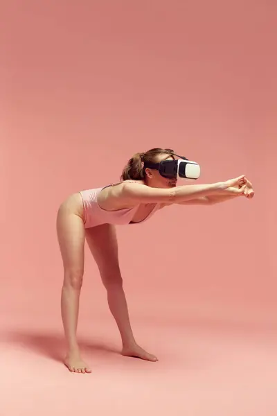 Virtual training. Woman in bodysuit, wearing vr glasses and doing stretching exercises against pink studio background. Concept of body and skin care, fitness, natural beauty, health, modern technology