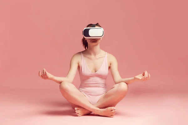 Online meditation and yoga. Woman in bodysuit, wearing vr glasses and sitting against pink studio background. Concept of body and skin care, fitness, natural beauty, health, modern technology
