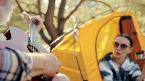 在温暖的日子里 一对年迈的恋人在森林里远足 女人坐在帐篷里喝茶听男人弹奏吉他闲暇时间 积极生活方式 业余爱好 自然的概念 — 图库视频影像