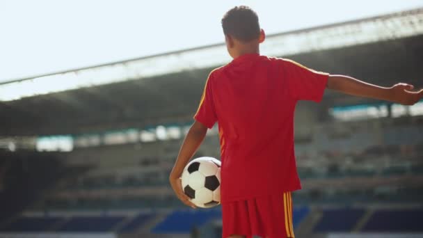 小男孩 身穿红色制服的孩子站在空荡荡的体育场 手拿着足球 梦想着成为一名足球运动员 想象和未来 体育的概念 — 图库视频影像