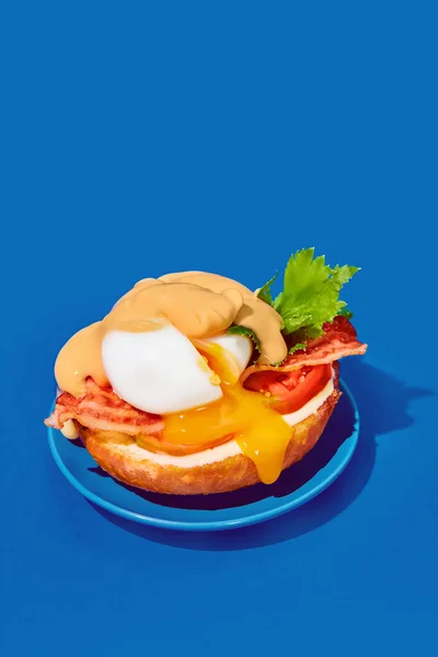 Poached eggs with liquid yolk, tomato, bacon and cheese sauce on toasted bun on plate on blue background. Concept of breakfast, food, taste, creativity. Pop art photography. Poster. Copy space for ad